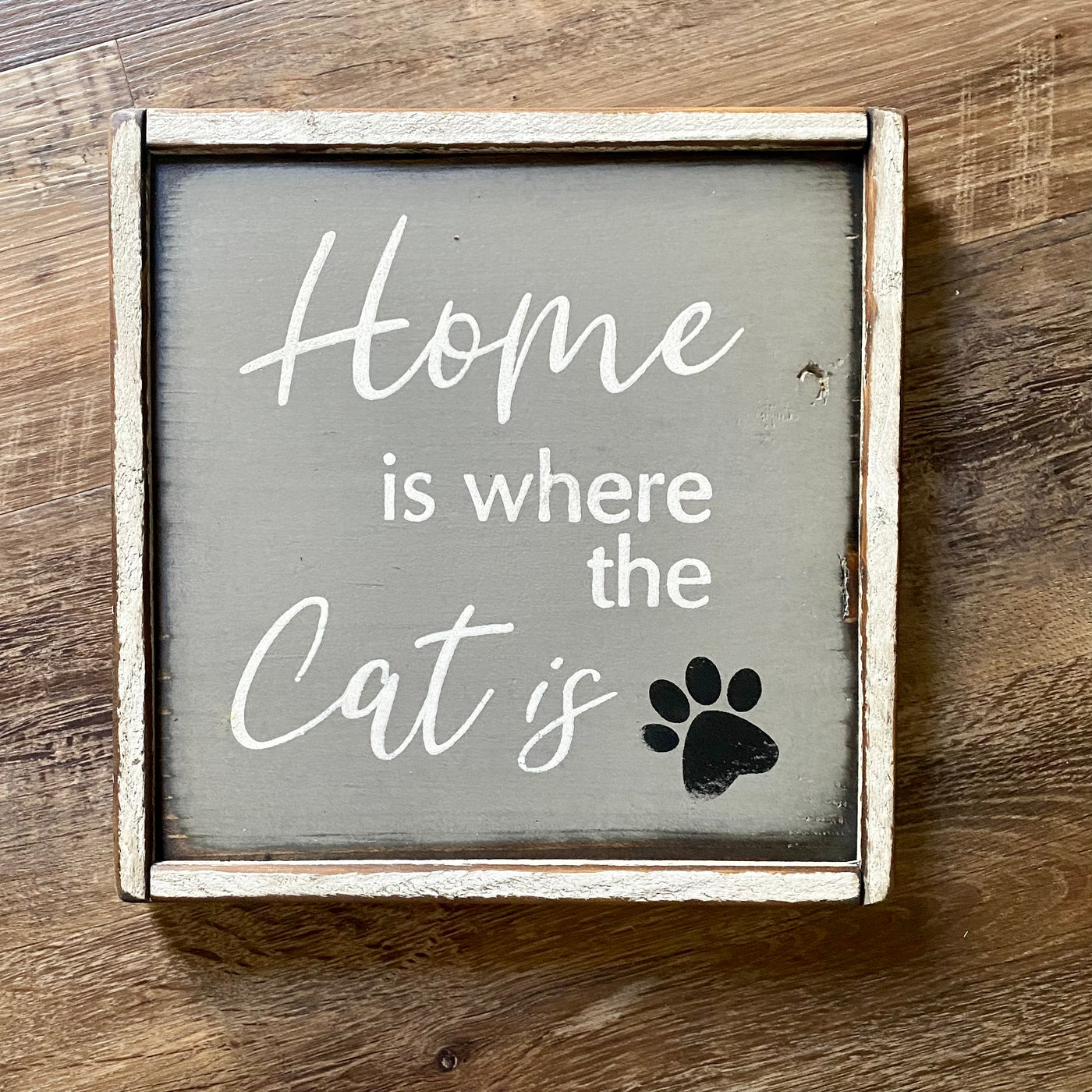 10x10 white framed wood sign reads home is where the cat is. Grey background with white lettering and a black pawprint accent.