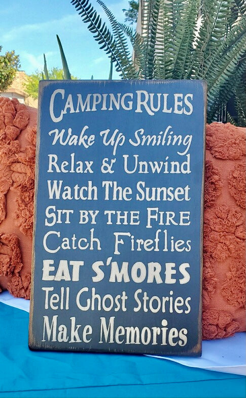 Wood sign reads camping rules, wake up smiling, relax & unwind, watch the sunset, sit by the fire, catch fireflies, eat s’mores, tell ghost stories, make memories. 