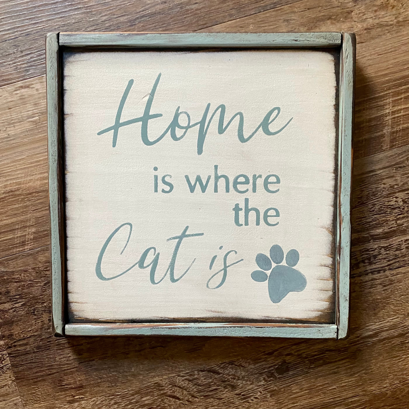 10x10 light blue framed wood sign reads home is where the cat is. White background light blue lettering.