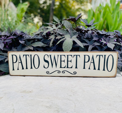 White wooden sign with natural edge reads Patio Sweet Patio in black lettering with a black decorative accent underneath wording